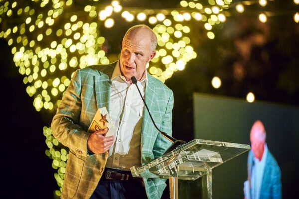 River Hero nominee Phil Wilson in a plaid jacket addressing the crowd from the podium beneath sparkling tree lights