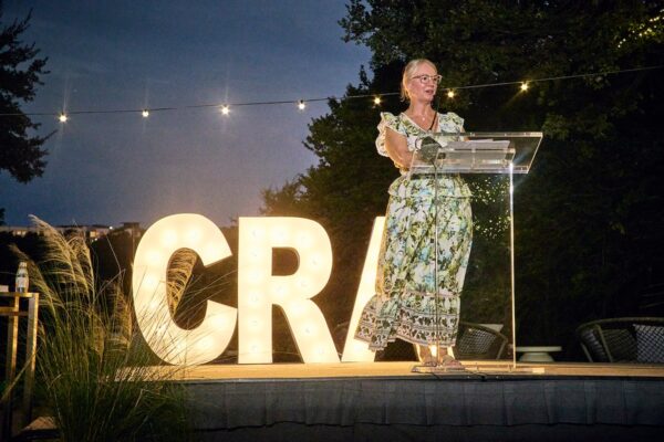 Executive Director Adrienne Longenecker speaking at a clear podium beside illuminated CRA marquee letters at night